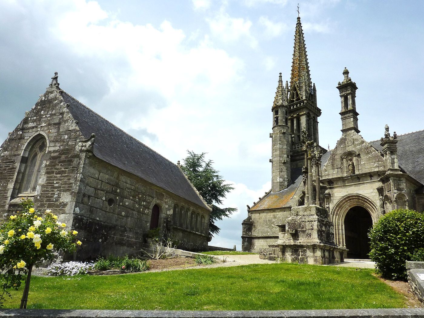 L'enclos paroissial de Brasparts : l'église, le calvaire et l'ossuaire en pierre grise, sous un ciel de nuages clairs