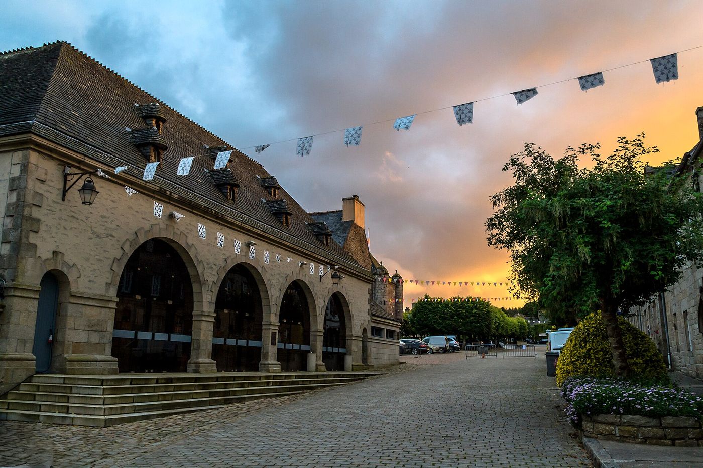Les halles de Guerlesquin, bâtisse de granite aux arcades, sous un ciel orangé et des fanions