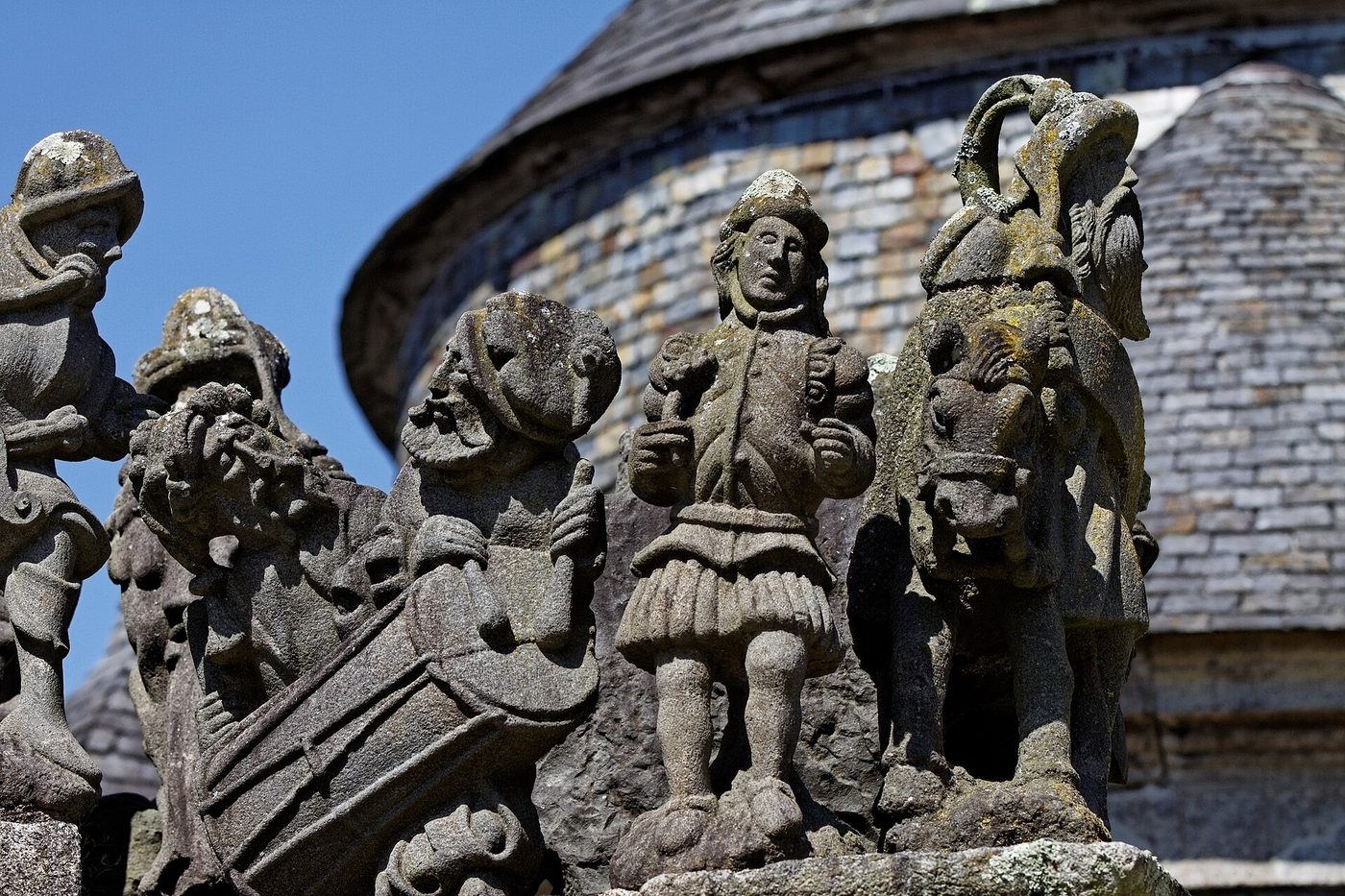 Personnages sculptés dans le granit sur le calvaire de l'enclos paroissial de Guimiliau, devant le toit d'ardoise de la chapelle