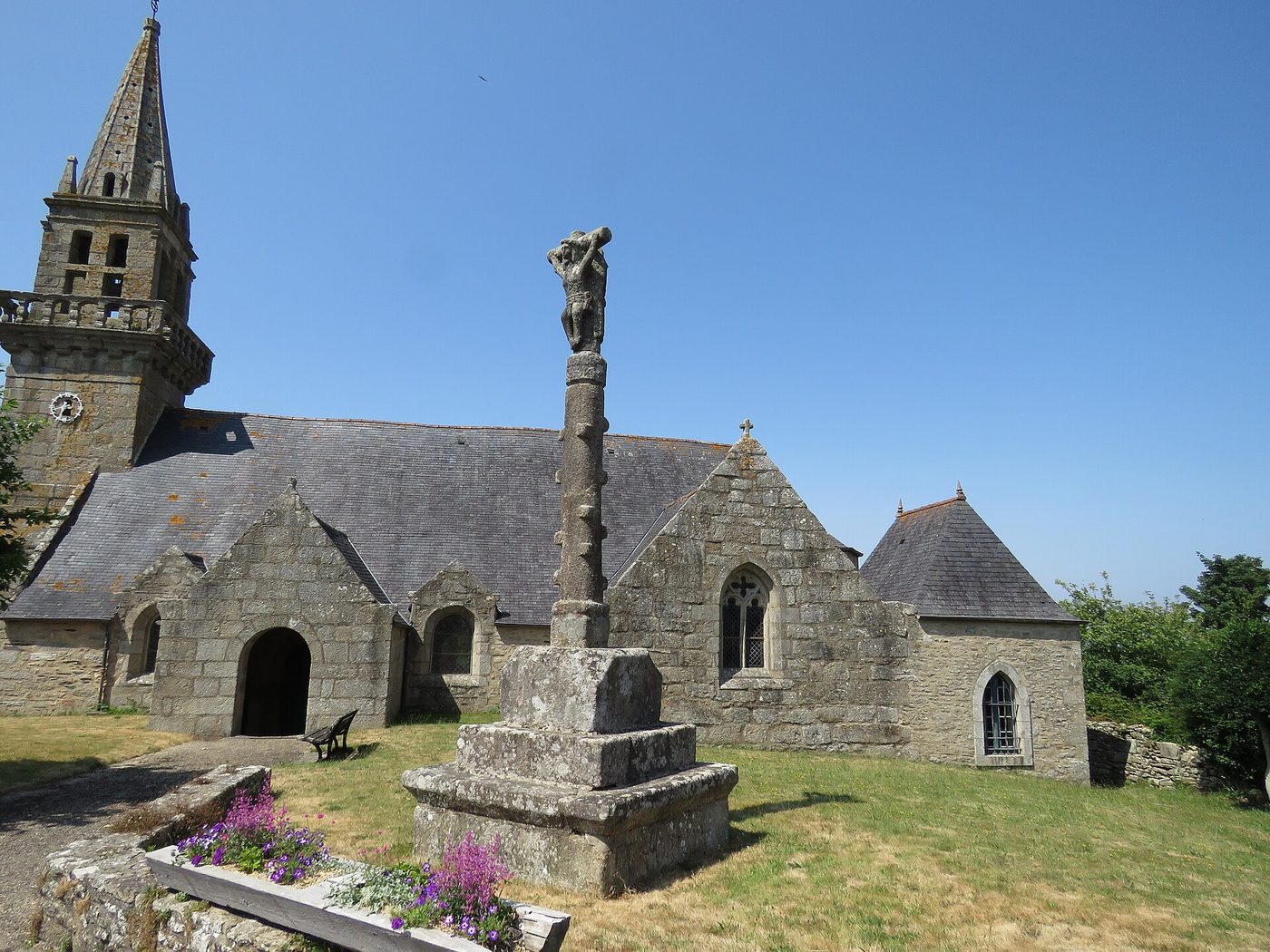 Église Notre-Dame du Cloître-Saint-Thégonnec, son clocher et la croix de pierre du calvaire sous un ciel bleu